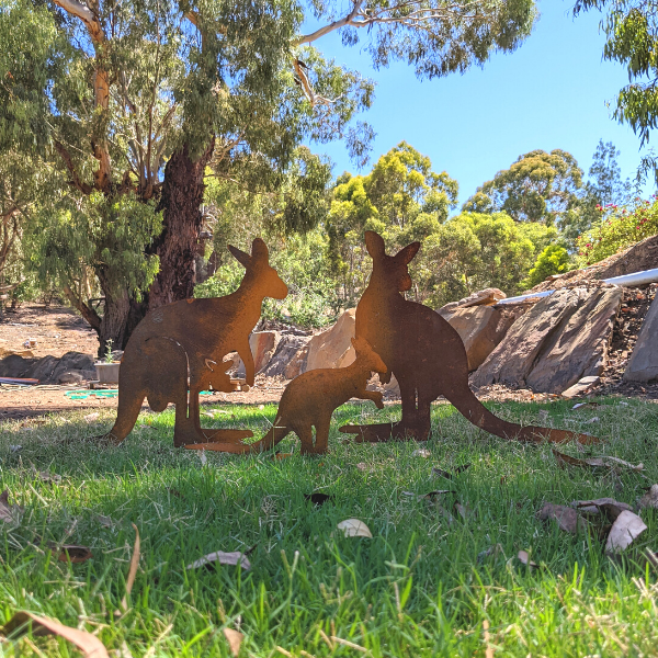 A family of 3 rusty metal kangaroos staked on lawn in a backyard. There are 3 separate metal cutouts: one kangaroo with a joey in her pouch, another adult kangaroo, and a small joey.