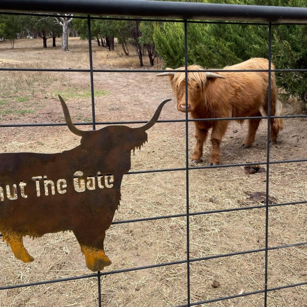 Image is a rusty metal sign in the silhouette of a Highland Cow with the words 'Shut the Gate', mounted on a stock gate. A live highland cow is behind the gate.