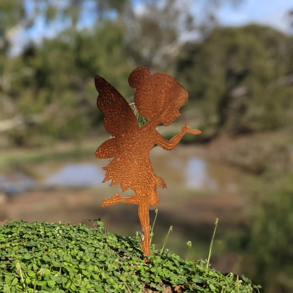 Image of a rusty metal fairy garden cutout, looking a little like tinkerbell, blowing a kiss standing outdoors in a patch of grass.  Background is blurred.