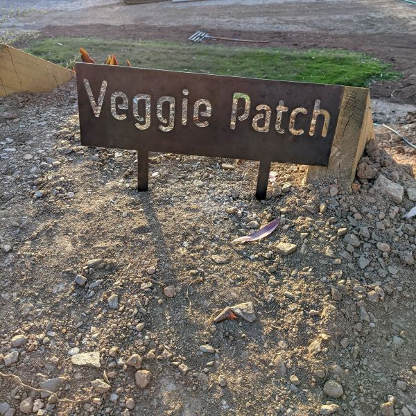 Image is a rectangular metal sign staked into the ground with the phrase "Veggie Patch" cut out.